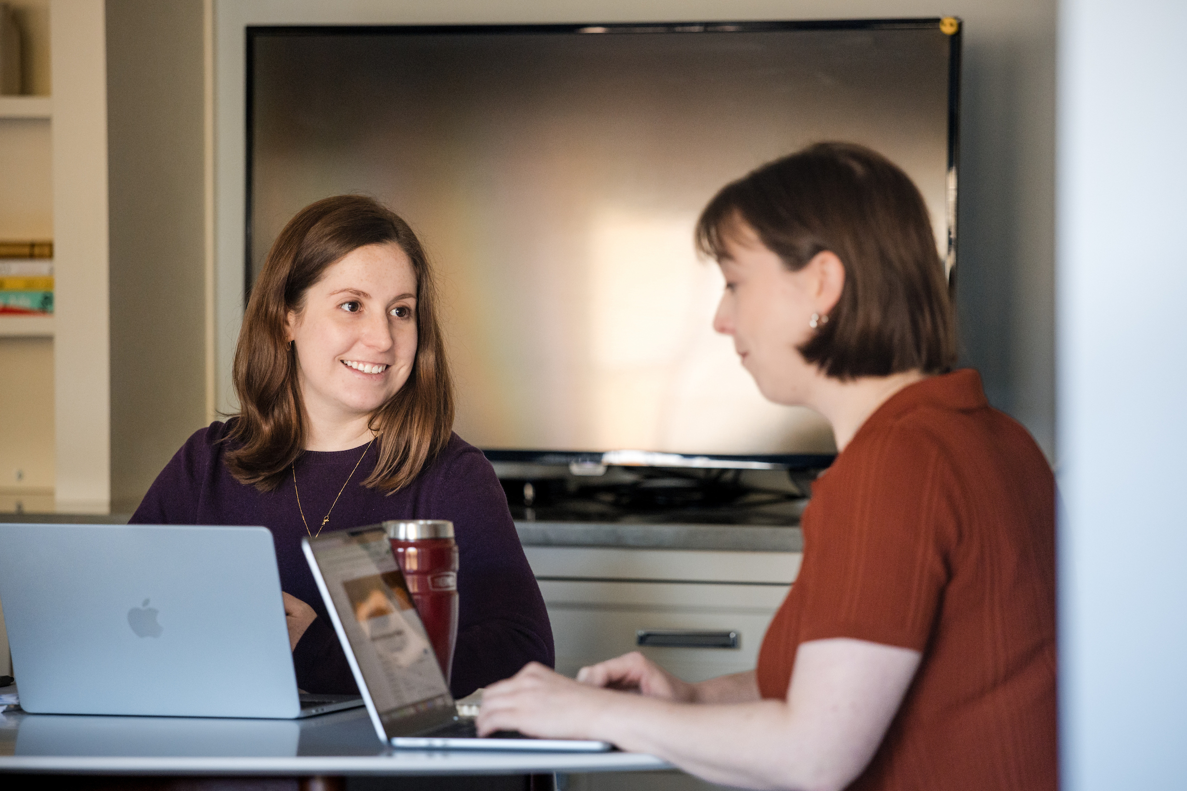 B Corp - A colleague smiles toward her coworker across two laptops, with the meeting room screen behind them.