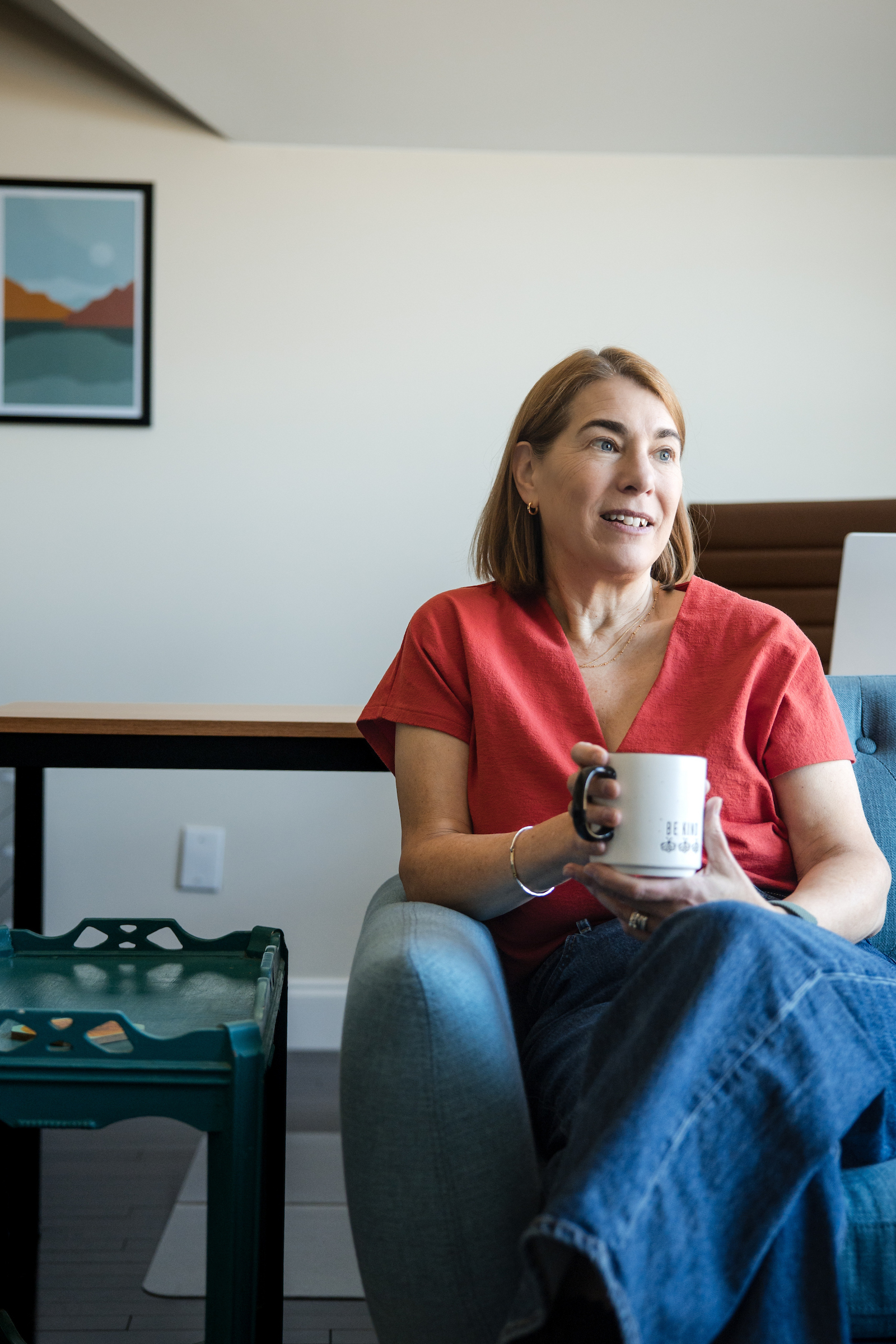 B Corp - Seated on the blue sofa, a colleague holds a mug and smiles toward someone outside the frame.